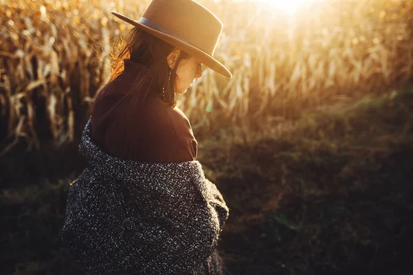 秋のフィールドで日没の光の中で歩く茶色の帽子とヴィンテージコートの美しいスタイリッシュな女性 大気中の瞬間 夕方田舎のトウモロコシ畑を歩くファッショナブルな若いヒップスター女性 — ストック写真