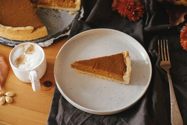 Pumpkin pie slice on modern plate and hot coffee on rustic table with linen napkin, autumn flowers and leaves. Homemade pumpkin tart recipe. Happy Thanksgiving