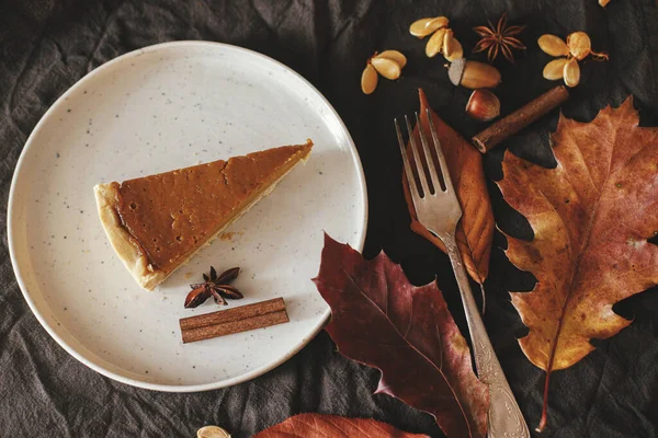 Pumpkin pie slice on modern plate on rustic table with linen napkin, autumn flowers and leaves, anise and cinnamon. Top view. Happy Thanksgiving. Homemade pumpkin tart recipe