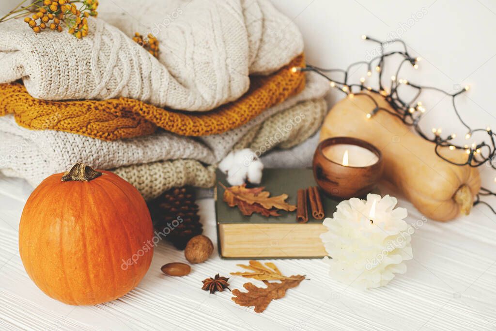 Cozy autumn days, slow living. Pumpkin, cozy sweaters, autumn leaves, burning candle and vintage book on white wooden background in room.Happy Thanksgiving