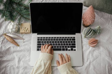 Eller dizüstü bilgisayarda, arka planda siyah ekranla modern Noel süsleri ve çam dalı olan şık bir yatağın üzerinde çalışıyor. Tatillerde serbest ve uzaktan çalışma. Yukarıya bak. Metin için boşluk