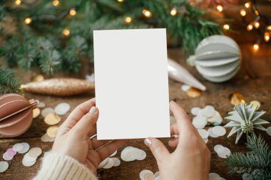 Ellerinde boş tebrik kartı, noel süsleri, konfeti, çam dalları ve kırsal ahşap arka planda ışıklar var. Noel kartı maketi. Mesaj için yer var. Mevsimler selamlar. Şablon