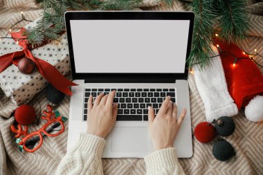 İnternetten alışveriş. Eller dizüstü bilgisayarda, boş ekranla, rahat yatakta, şık Noel süsleri, hediye ve Noel Baba şapkası ile çalışıyor. Serbest çalışma ve tatillerde uzaktan çalışma. Mesaj için yer var. Üst yaşam