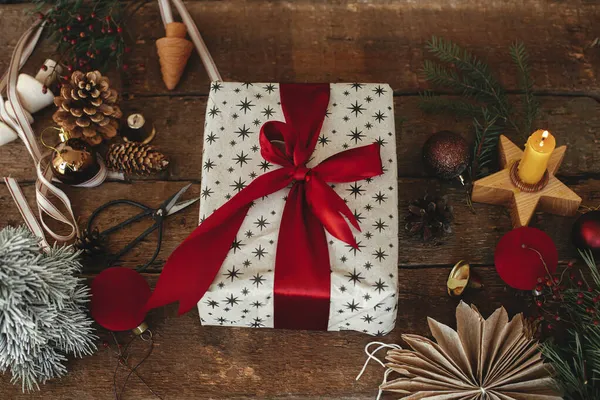 Kırsal ahşap masa üzerinde kırmızı kurdeleyle süslenmiş şık bir Noel hediyesi. Bayram süsleri, üst manzara. Xmas hediye ve takılar, mum, makas, kozalak, köknar dalları, yıldız