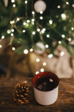Tarz bardağı ve çam kozalağı kırsal ahşap üzerinde akşam odasındaki ışıklarda Noel ağacının arka planında. Mesaj için yer var. Dekoratif odada çay ile seramik bardak. Atmosferik Noel zamanı