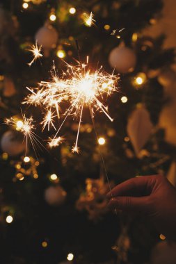 Kutlama odasındaki Noel ağacı ışıklarının arka planında yanan bir maytap tutuyor. Mutlu yıllar! Kadın elinde parlayan havai fişek bengi. Mesaj için yer var. Atmosferik an