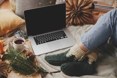 Diz üstü bilgisayarı, Noel ışıkları, çay, yıldızlar, çam, mum ve yastıklı yumuşak çoraplı kadın ayakları şık bir odada. Evdeki sıcak kış tatili. Serbest ve günlük