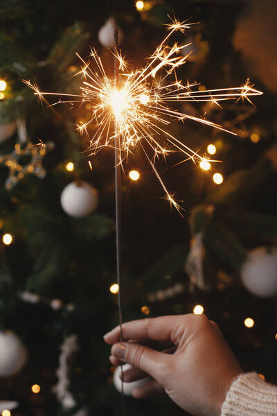 Happy New Year! Hand holding burning sparkler on background of christmas tree lights in festive room. Firework bengal glowing in woman hand. Space for text. Atmospheric moment. Merry christmas