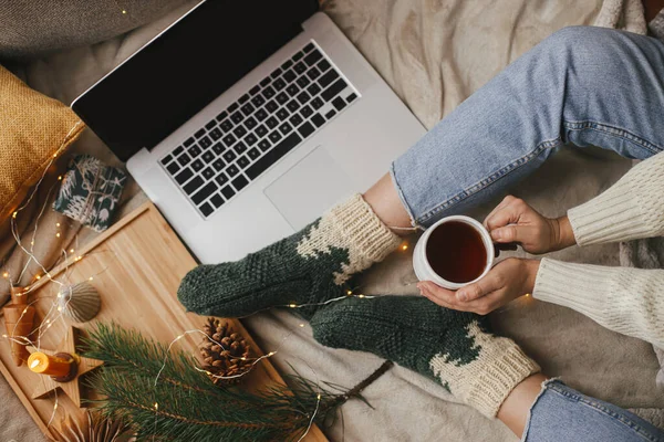 Evdeki sıcak kış tatili. Sıcak bir fincan çayı olan kadın yumuşak yatağında dizüstü bilgisayarla, noel ışıklarıyla, yıldızlarla, çam ağacıyla, mumla ve şık bir odada yastıklarla dinleniyor. Serbest çalışmak ve blog yazmak. Üst görünüm