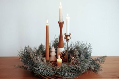 Noel yaklaşıyor. Noel çelenginde yanan şık mumlar, çam kozalakları ve modern festival odasındaki beyaz duvarın arka planındaki ahşap masa üzerindeki ağaç dekoru. Atmosferik kış tatili