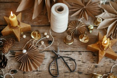 Şık Noel çanları, kağıt melek ve yıldızlar, kozalaklar, mum, iplik ve makas kırsal ahşap arka planda. Şenlikli İskandinav çelengi yapmak, karamsar bir görüntü. El yapımı tatil dekorasyonu