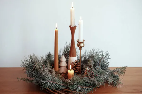 圣诞节来了在现代喜庆的房间里 圣诞花圈里燃烧着时尚的蜡烛 花圈上有松果和装饰在白色墙壁后面的木桌上的树木 大气冬季假期 — 图库照片