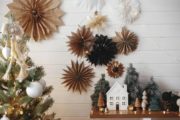 メリークリスマス 白い木製の壁に紙の星の背景にスタイリッシュなクリスマスの小さな家 黄金のライト 現代のスカンディナヴィアの部屋で大気中のお祝いの装飾 魔法の冬時間 — ストック写真