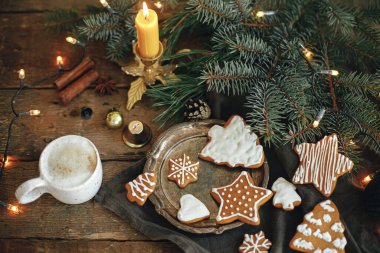 Noel kurabiyeleri, şık beyaz bardakta kahve, köknar dalları, peçetedeki sıcak ışıklar ve kırsal ahşap masa. Atmosferik görüntü. Kışın kırsal kesim. İyi tatiller.!