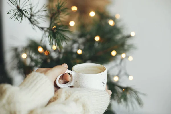 Hands in cozy sweater holding warm coffee in stylish cup on background of fir branches with warm lights in festive scandinavian room. Atmospheric winter time and hygge home. Merry Christmas!
