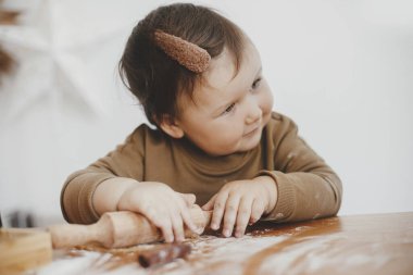 Kurabiyeler için ahşap broşla hamur yoğuran sevimli bir bebek. Küçük tatlı kızın annesine Noel kurabiyesi yapmada yardım ediyor. Otantik güzel anlar