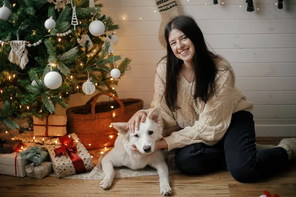 クリスマスツリーの下にかわいい犬とギフトやライトと座って居心地の良いセーターでスタイリッシュな幸せな女性 お祝いのスカンディナヴィアの部屋でかわいい犬を愛撫若い女性 ペットと冬の休日 — ストック写真