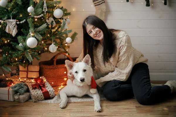 クリスマスツリーの下にかわいい犬とギフトやライトと座って居心地の良いセーターでスタイリッシュな幸せな女性 お祝いのスカンディナヴィアの部屋でかわいい犬を愛撫若い女性 ペットと冬の休日 — ストック写真