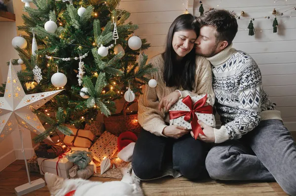 メリークリスマス クリスマスツリーを背景にライトで赤い弓とお祝いの装飾された部屋でかわいい犬を持つスタイリッシュなカップル 現在を受け入れ交換する若い家族 — ストック写真