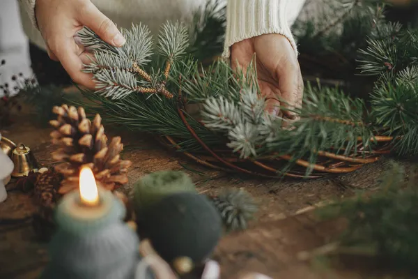 Making rustic christmas wreath close up. Woman hands holding fir branch and arranging christmas wreath on rustic wooden background with candle, pine cones, thread. Winter holiday preparations