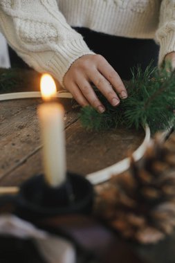 Eller, köknar dalları, kurdeleler, yuvarlak ahşap halka, makas, iplik, mum ile kırsal masada modern Noel çelengi yapıyor. Atmosferik karamsar görüntü. Kış tatili hazırlığı