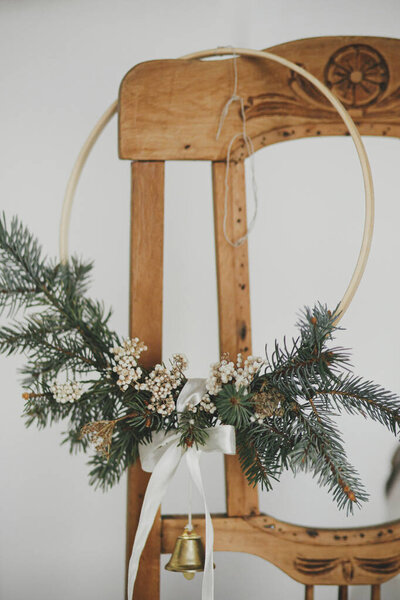 Merry Christmas! Modern christmas wreath with bell hanging on rustic wooden chair. Winter holidays preparation, atmospheric moody image. Stylish boho xmas wreath in scandinavian room