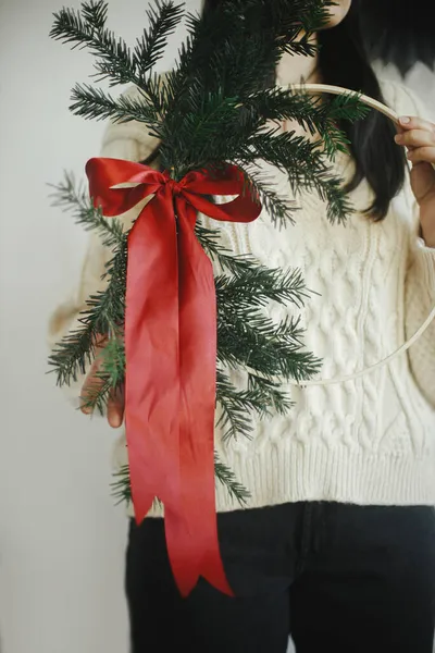 白い壁の背景にモダンなクリスマスリースを保持居心地の良いセーターでスタイリッシュな女性 メリークリスマスとハッピーホリデー モミの枝と手に赤い弓でスタイリッシュなクリスマスリース — ストック写真