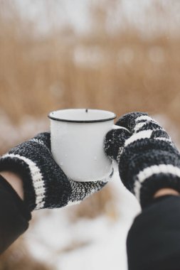 Elleri rahat eldivenlerde, kışın kurumuş çimlerin ve kar gölünün arka planında sıcak bir fincan çay tutuyor. Soğuk kış mevsiminde yürüyüş ve seyahat. Sıcak bir içecek. Metin için boşluk