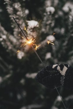 Mutlu yıllar! Karda çam ağacı dallarının arka planında yanan havai fişeklerle sıcak bir eldiven tutan kadın. Parlayan maytapla el ele tutuşmak. Atmosferik sihirli an