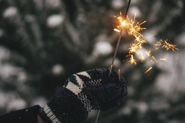 Karda çam ağacı dallarının arka planında yanan maytap tutan sıcak eldiven. Mutlu yıllar! Atmosferik sihir anı. Akşamları elinde parlayan havai fişeklerle kadın eli. İyi tatiller.!