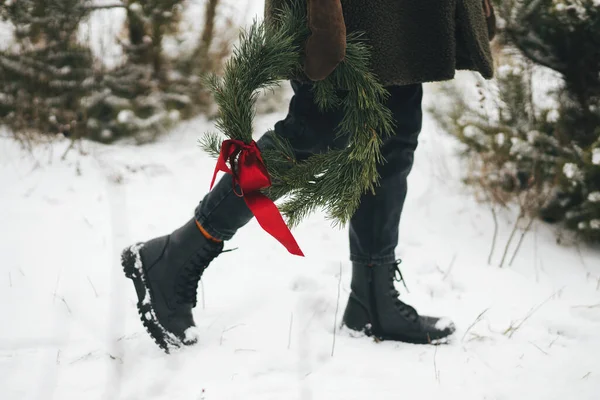 田舎の冬休み 冬の公園でクリスマスリースとスタイリッシュな女性 若いです女性で居心地の良いミトンとコートを保持Xmas花輪で雪の冬のフィールドで赤い弓 クロップドビュー — ストック写真