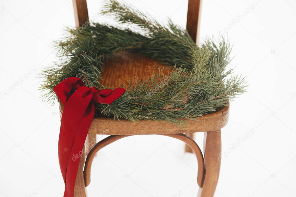 Merry Christmas! Christmas wreath on rustic chair in snowy winter field. Winter holidays in countryside. Stylish xmas wreath with pine branches and red bow on wooden chair. Atmospheric time