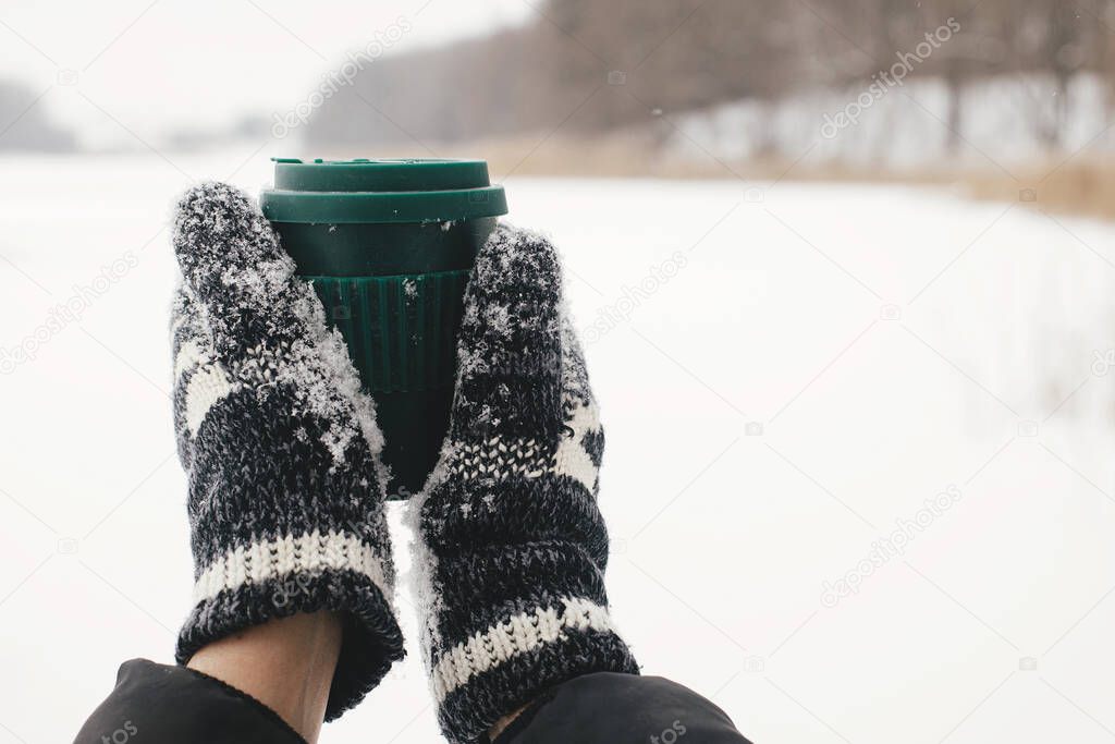 Plastic free cup with warm drink. Hands in cozy gloves holding reusable warm cup of tea on background of snow lake in winter. Hiking and traveling in cold winter season. Sustainable lifestyle