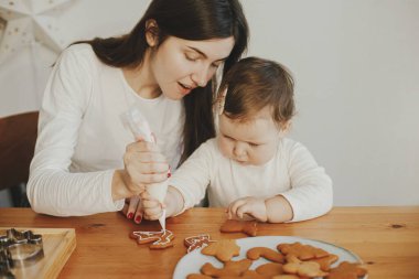 Sevimli küçük kız ve anne, Noel kurabiyelerini modern odadaki ahşap masada şekerle süslüyor. Mutlu aile zamanları, tatil hazırlıkları. İyi tatiller.!