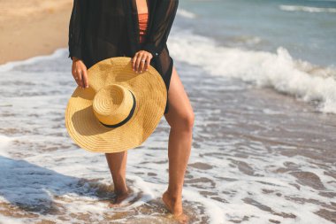 Summer vacation. Carefree woman holding hat on sunny beach at sea waves, close up. Stylish young fit female with straw hat in hands relaxing on tropical shore. Space for text