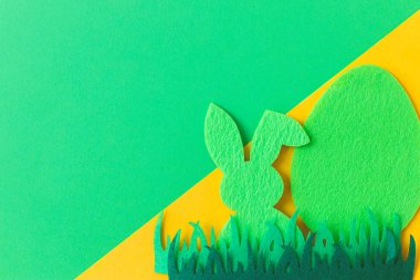 Green Easter egg and bunny in grass on green and yellow background, flat lay with space for text. Happy Easter! Colorful artificial egg and bunny decor. Creative Easter hunt concept