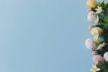 Happy Easter! Colorful Easter chocolate eggs and spring flowers border on blue background, flat lay with space for text. Stylish easter greeting card template