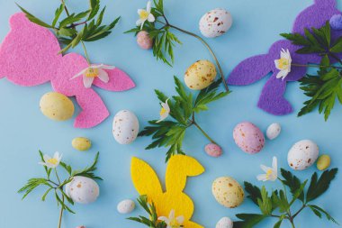 Happy Easter! Colorful Easter bunnies, chocolate eggs and spring flowers on blue background flat lay. Pink and yellow artificial decor and fresh spring flowers. Easter hunt