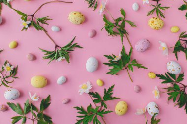 Happy Easter!  Colorful Easter chocolate eggs and spring flowers flat lay on pink background. Stylish easter layout. Greeting card or banner. Candy and flowers composition
