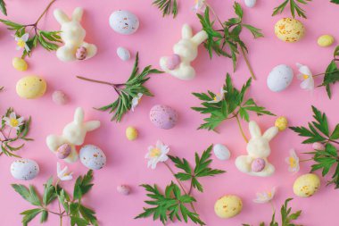 Colorful Easter chocolate eggs, bunnies and spring flowers flat lay on pink background. Happy Easter! Stylish easter layout. Greeting card or banner. Candy and flowers composition