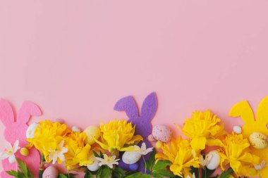 Mutlu Paskalyalar! Renkli Paskalya tavşanları, çikolatalı yumurtalar ve nergisler pembe arka planda sınırda, düz bir şekilde uzanıyorlar. Pembe ve sarı yapay dekor ve bahar çiçekleri. Paskalya avı
