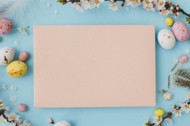 Stylish empty card, colorful Easter chocolate eggs, cherry blossoms, feathers on blue background flat lay. Easter greeting card mock up. Happy Easter! Postcard template