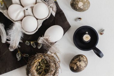 Paskalya kırsalının şık yatağı. Tepside doğal yumurtalar, tüyler, söğüt dalları, beyaz ahşap masadaki keten peçeteye yuva. Basit kırsal Paskalya natürmortu, pastel kahverengi renkte estetik.