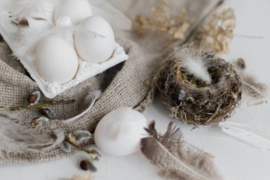 Paskalya kırsalında hayat. Tepside doğal paskalya yumurtaları, tüyler, söğüt dalları, beyaz ahşap masada çuval bezi üzerine yuva. Pastel bej renklerinde basit kırsal Paskalya estetiği. Mutlu Paskalyalar