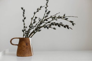 Willow branches in stylish rustic vase on wooden table. Simple Easter rural minimal aesthetics. Modern Easter still life. Pussy willow branches in home, natural spring decor. Space for text