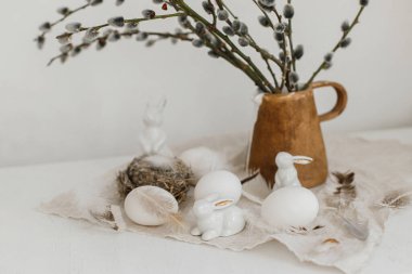 Mutlu Paskalyalar! Doğal yumurtalar, tavşan heykelcikler, tüyler, yuva, eski ahşap masadaki vazoda söğüt dalları. Paskalya kırsalında hayat. Masada basit bir paskalya süslemesi.