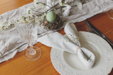 Paskalya brunch 'ı için şık bir ortam. Yuvada Paskalya yumurtası, tabakta tavşan peçetesi, çatal bıçak takımı, tavşan, bahar çiçekleri ve ahşap masada kırsal kumaş. Paskalya düzenleme ve çevre dostu dekorasyon