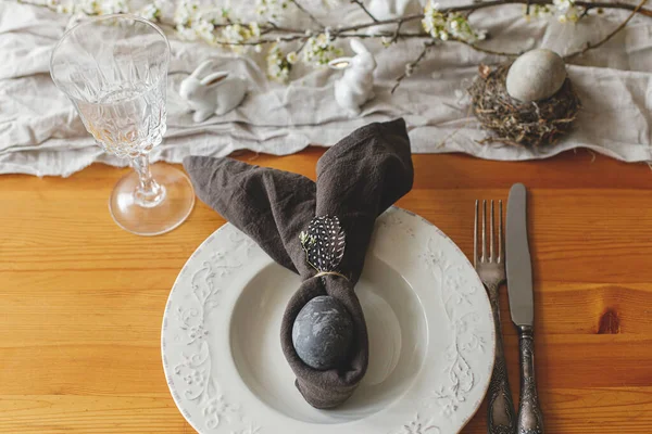 Stylish Easter brunch table setting. Easter egg in bunny napkin on plate with cutlery, bunny, spring flowers and rustic cloth on wooden table. Top view. Happy Easter!