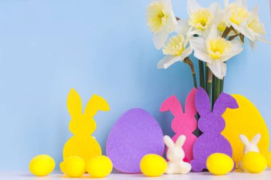 Mutlu Paskalyalar! Renkli Paskalya tavşanları, yumurtalar ve mavi arka planda nergis buketi ve metin için yer var. Pembe ve sarı yapay yumurta ve bahar çiçekli tavşan dekoru.