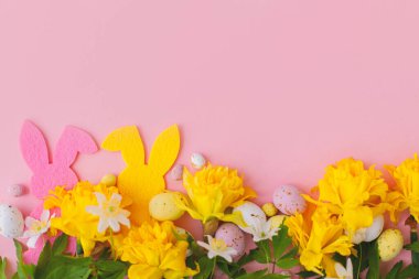 Mutlu Paskalyalar! Renkli Paskalya tavşanları, çikolatalı yumurtalar ve nergisler pembe arka planda sınırda, düz bir şekilde uzanıyorlar. Pembe ve sarı yapay dekor ve bahar çiçekleri. Paskalya avı